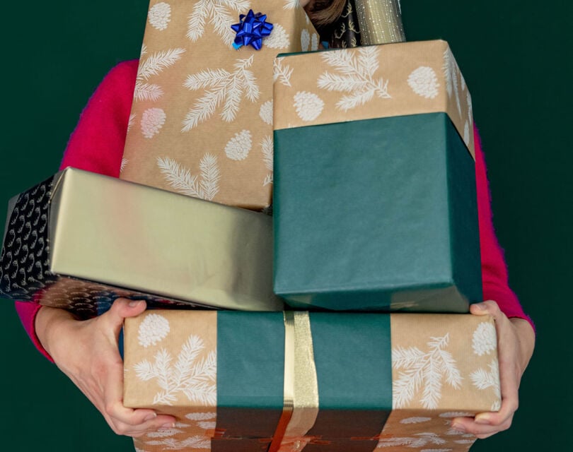 Een persoon balanceert 5 grote op elkaar gestapelde kerstcadeaus ingepakt in goud, groen en bruin inpakpapier