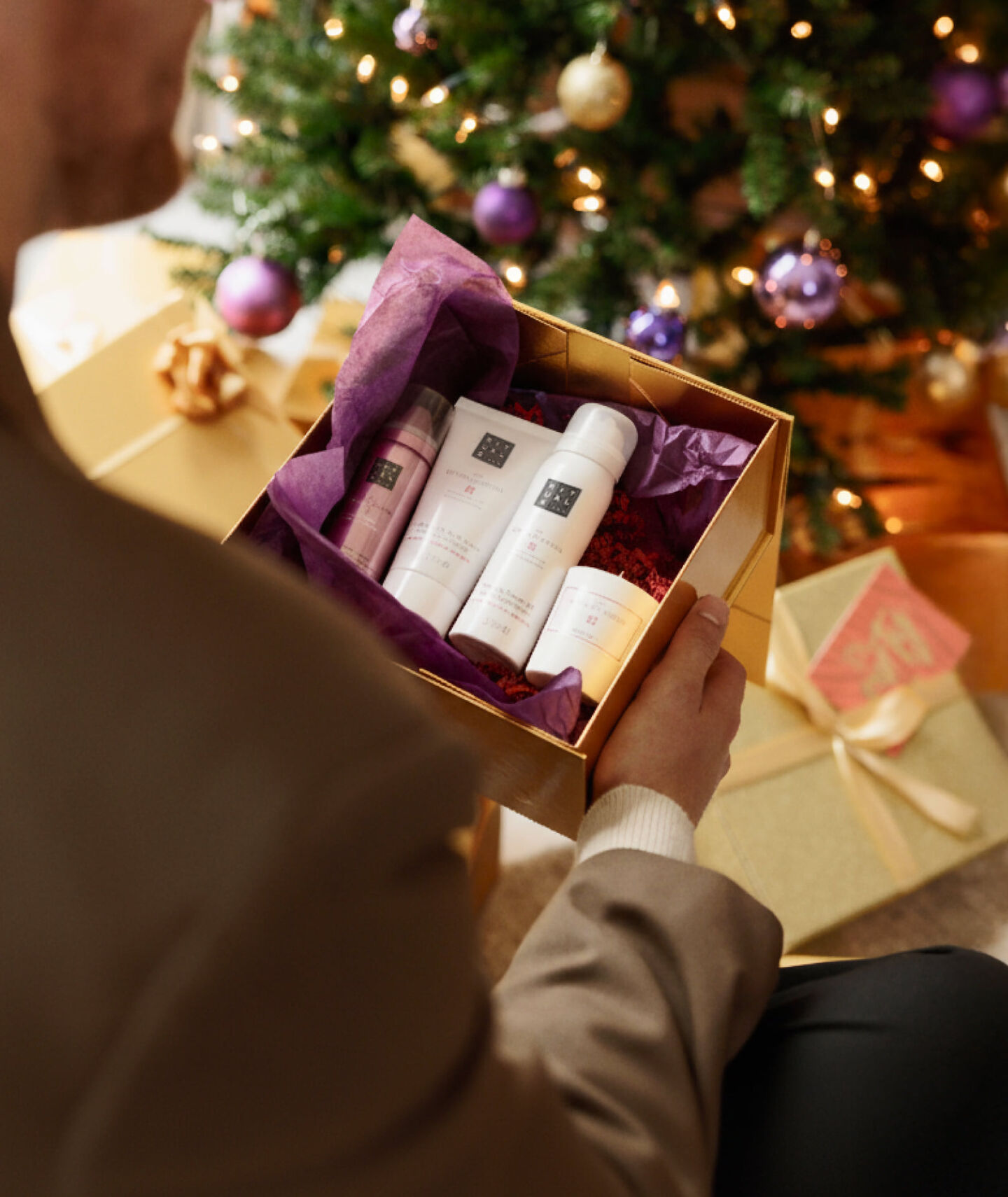 Iemand pakt een kerstcadeau met Rituals producten erin uit en op de achtergrond zie je een kerstboom met kerstpakjes onder