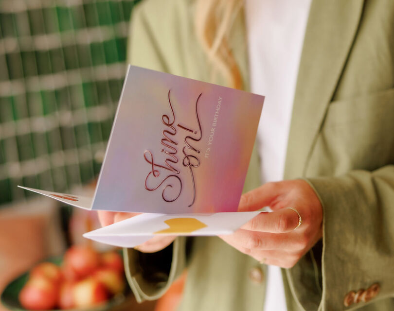 Een vrouw in een groene blazer houdt een kaart vast met de tekst 'Shine on''