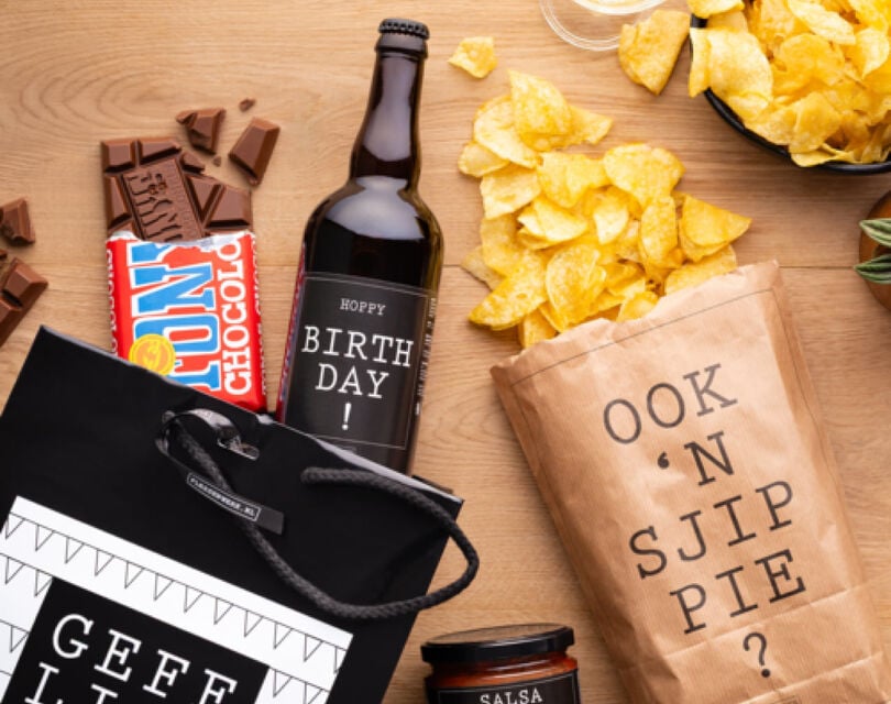 een assortiment aan verjaardagscadeaus ligt op een houten tafel met onder andere gepersonaliseerd bier, tony's chocolonely, en chips